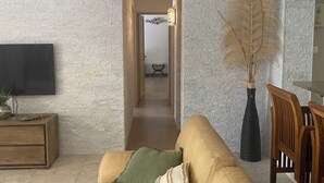 Un salon avec un canapé beige, une table basse en bois, un téléviseur fixé au mur et un vase contenant des plantes séchées.