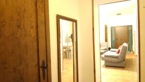 Ein Zimmer mit Holztür, einem Spiegel, der eine Person im Hintergrund reflektiert, und einem weißen Sofa mit rotem Kissen.