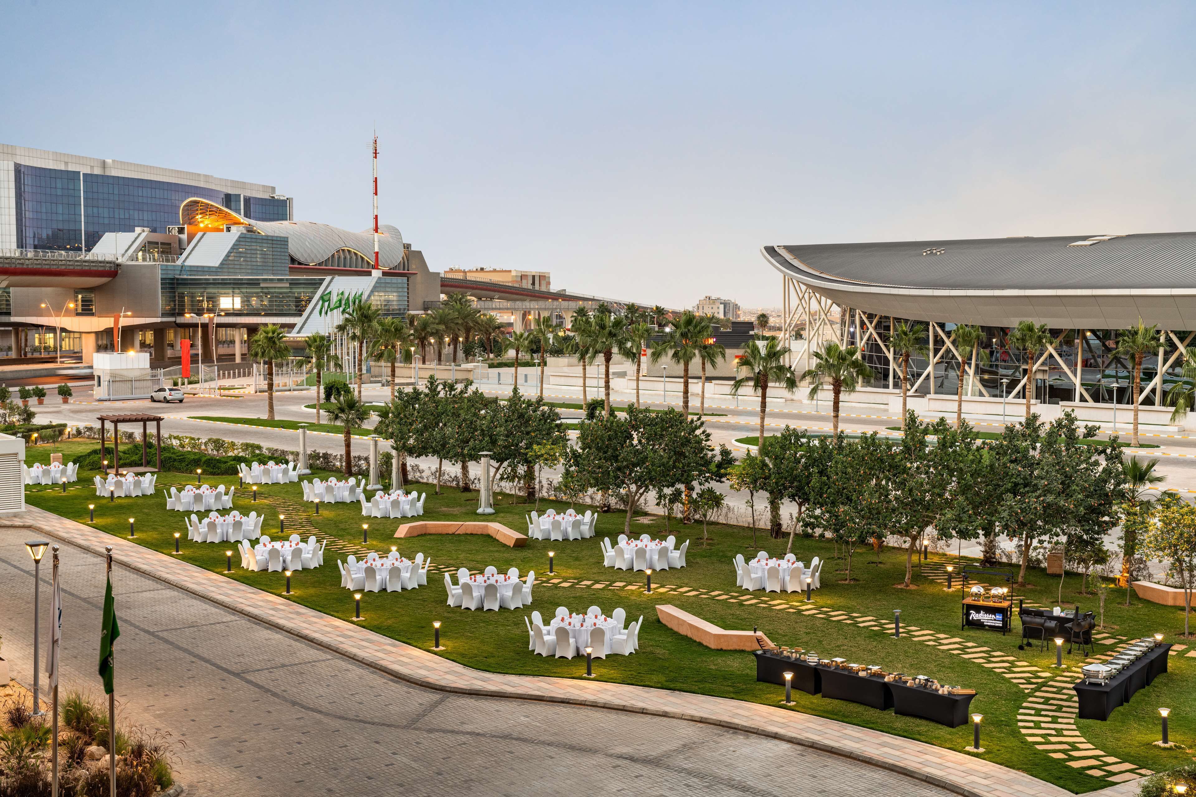 An outdoor event space with tables and chairs arranged on a lawn, surrounded by palm trees and a modern building in the background.