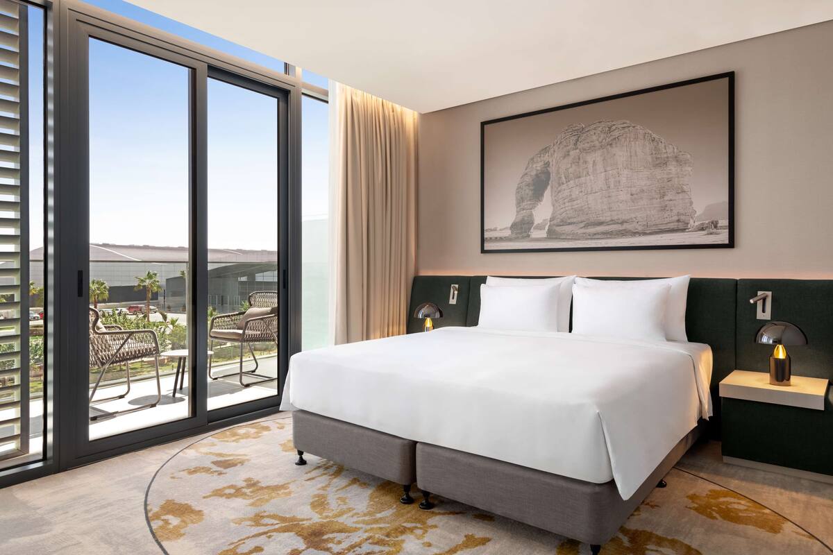 A modern hotel room with a large bed, a view through floor-to-ceiling windows, a framed artwork on the wall, and a small bedside table with a lamp.