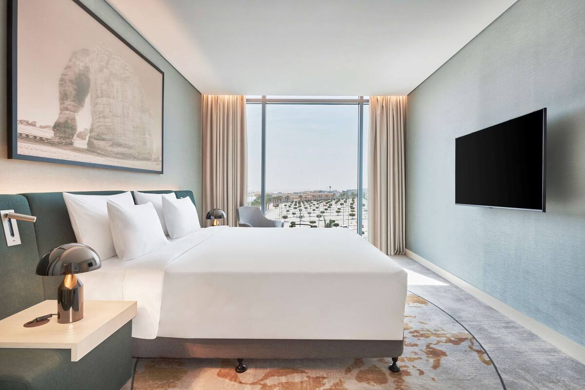 A modern hotel room with a large bed, a mounted TV, and a city view through large windows.