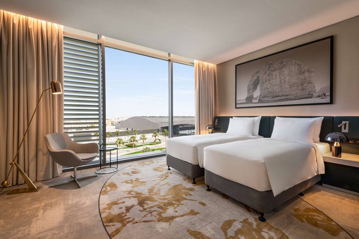 A hotel room with a large window, two beds, a chair, and a desk.