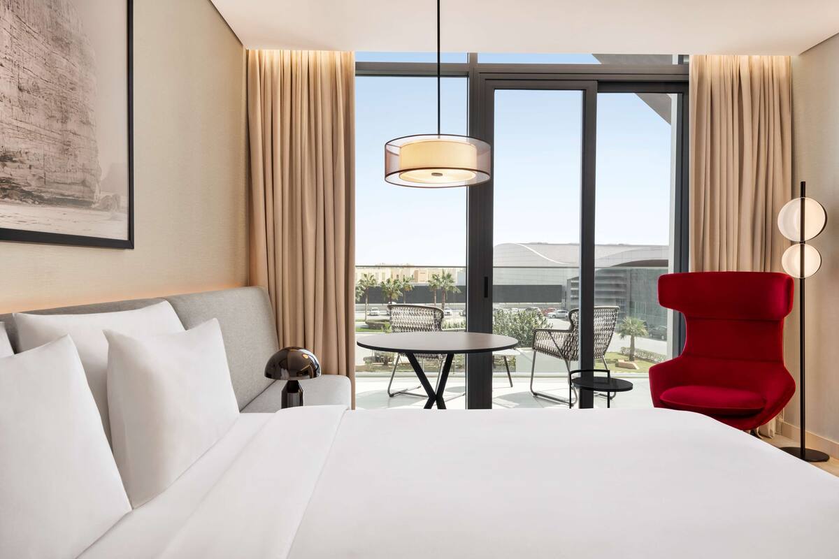 A hotel room with a large bed, a red chair, a small table, and a view of the cityscape through large windows.