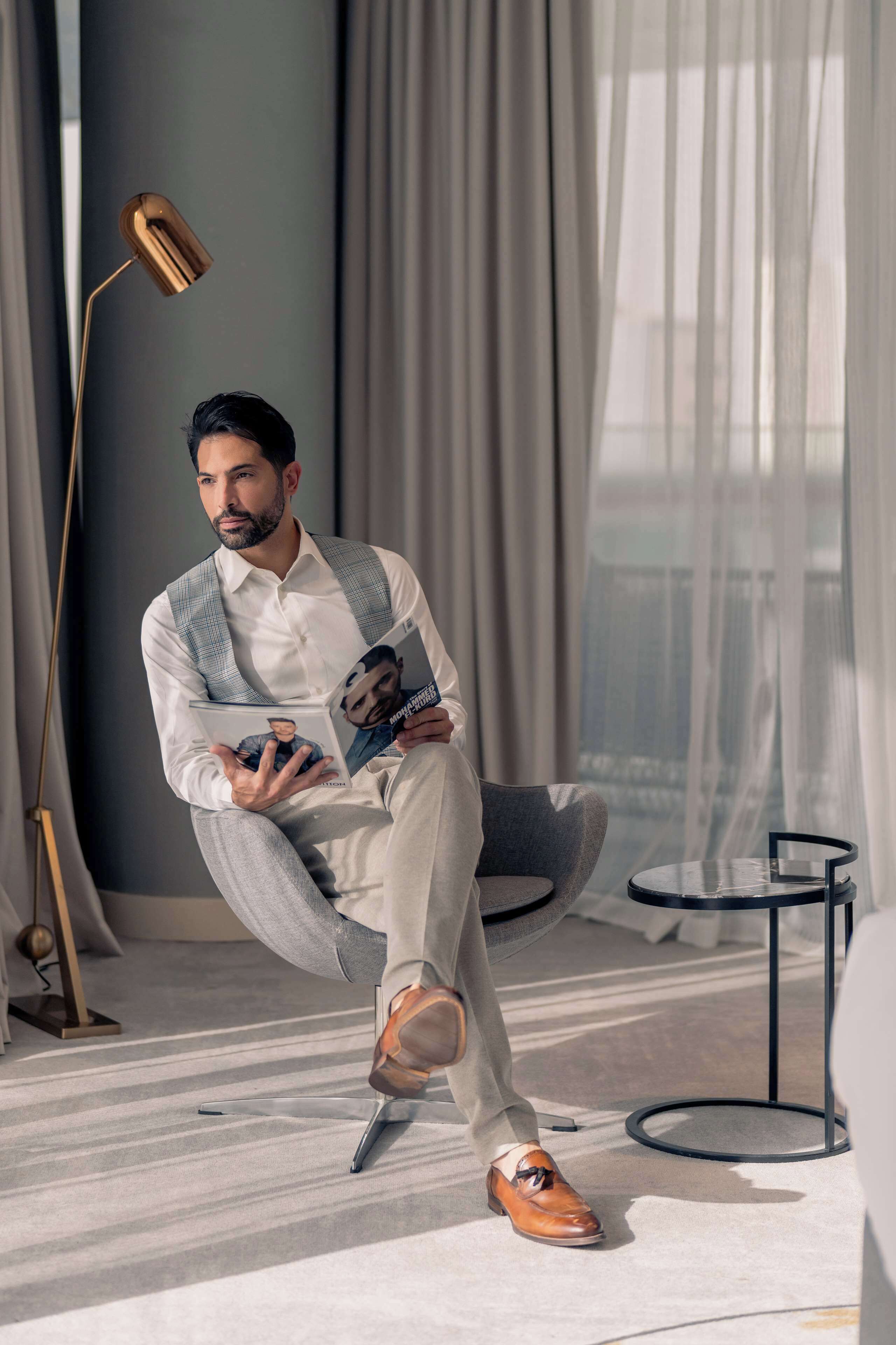 A man sitting on a chair in a modern room with a large window, a floor lamp, and a small round table.