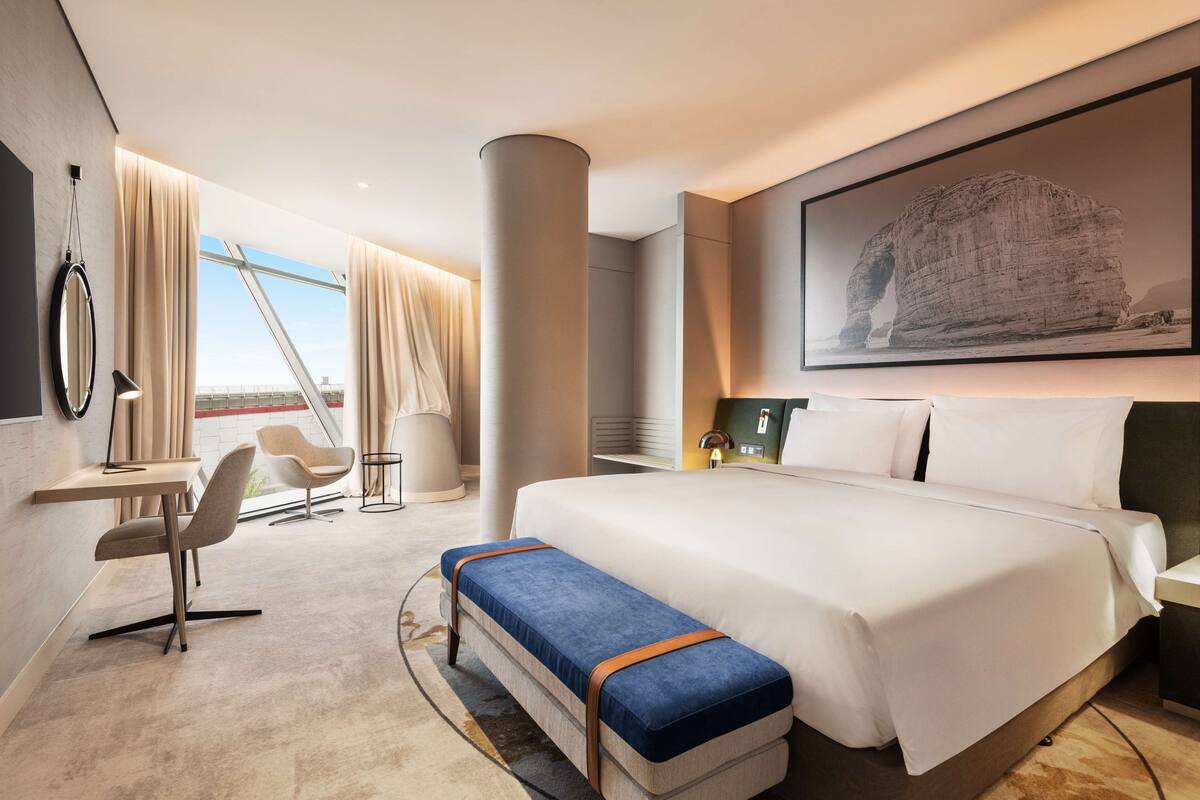 A modern hotel room with a large bed, a desk, a chair, and a view of the outdoors.