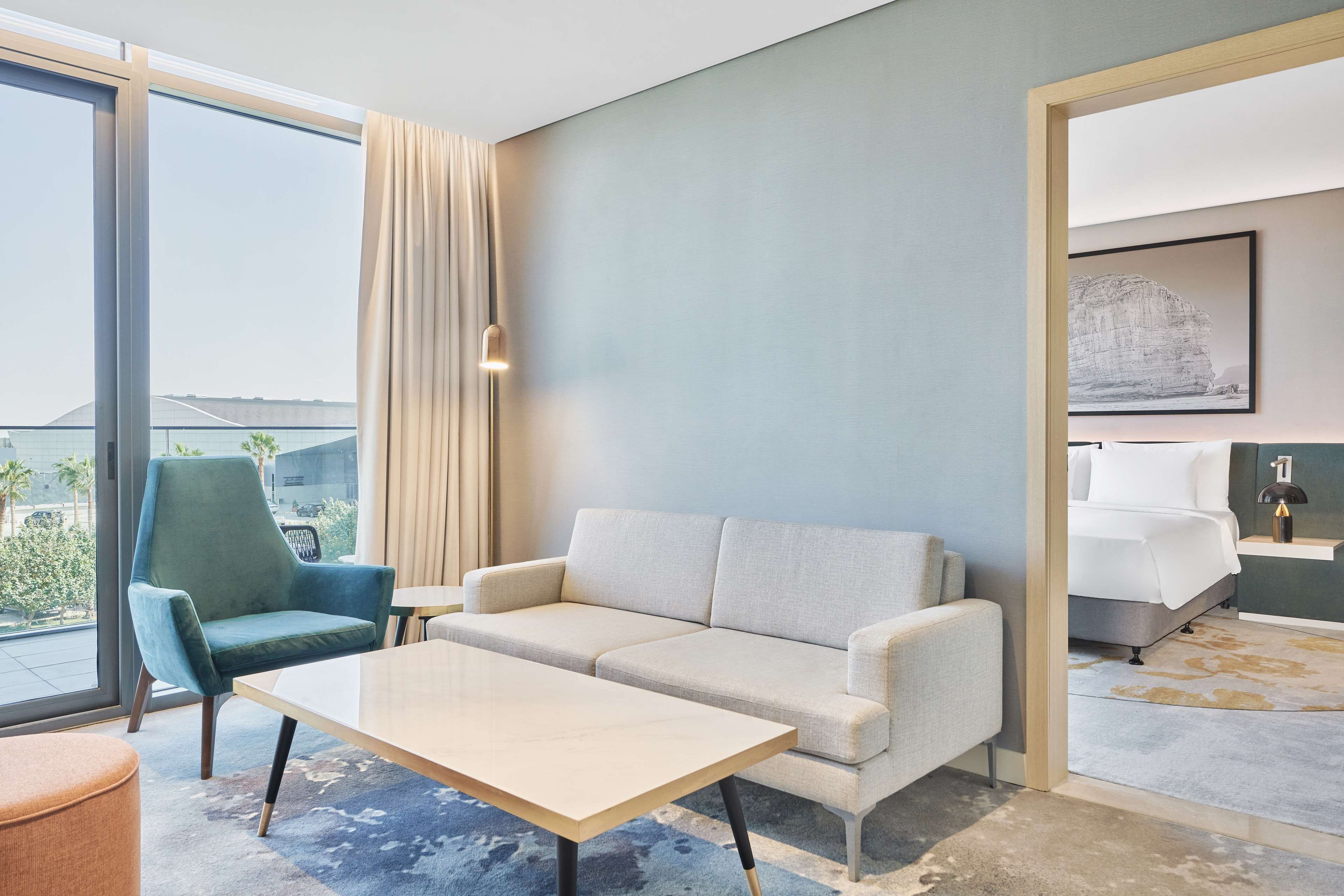 A modern hotel room with a large window, a sofa, a table, and a bed.