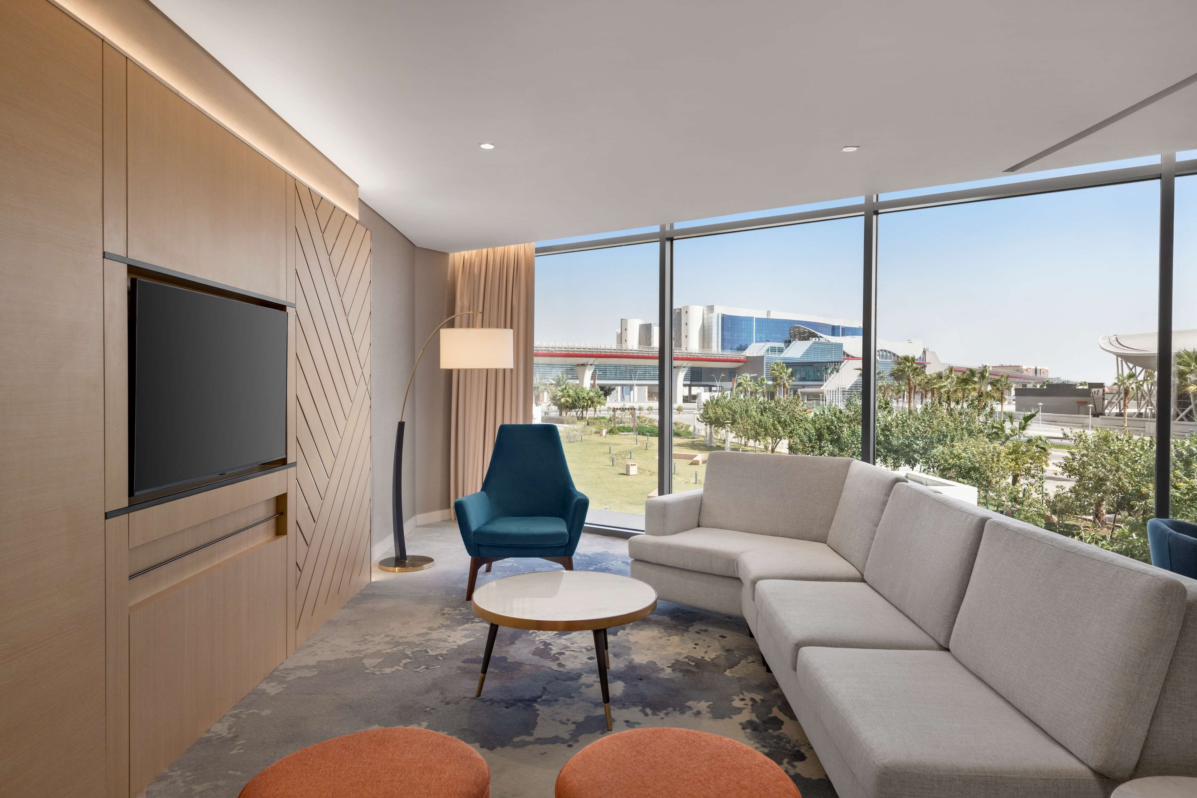 A modern hotel room with a large window, a flat-screen TV, a sofa, a chair, and a small table.