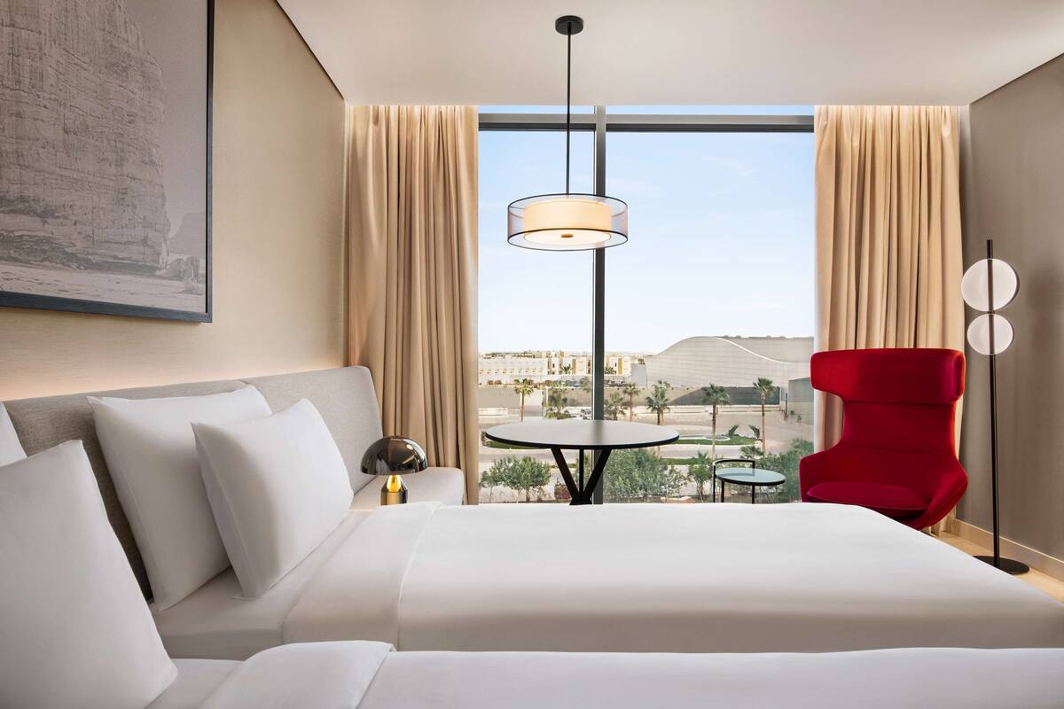 A hotel room with a large bed, a red chair, a small table, and a view of the city through large windows.