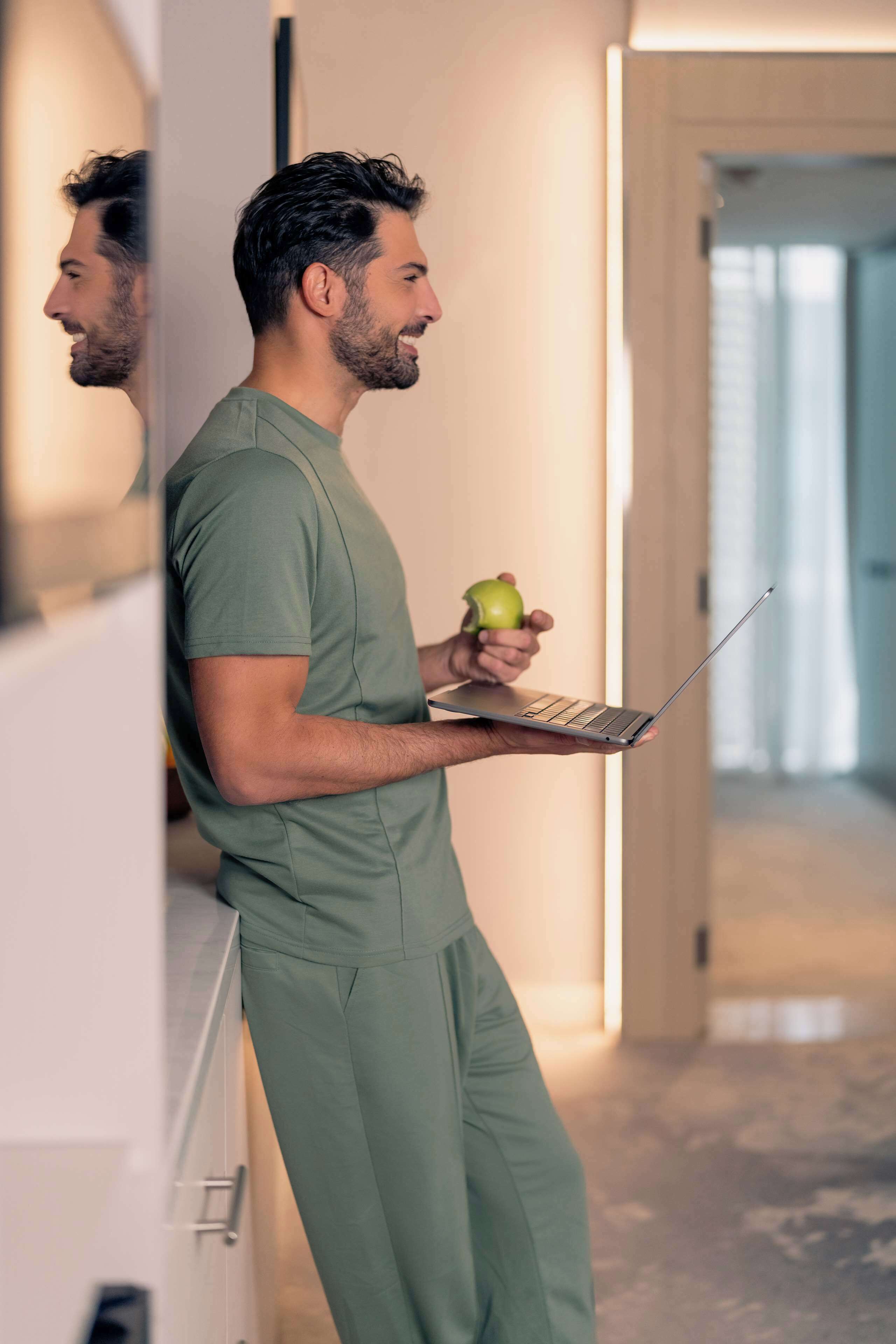 A person in a green uniform holding a laptop and an apple, standing in a hallway with a door and a light fixture in the background.