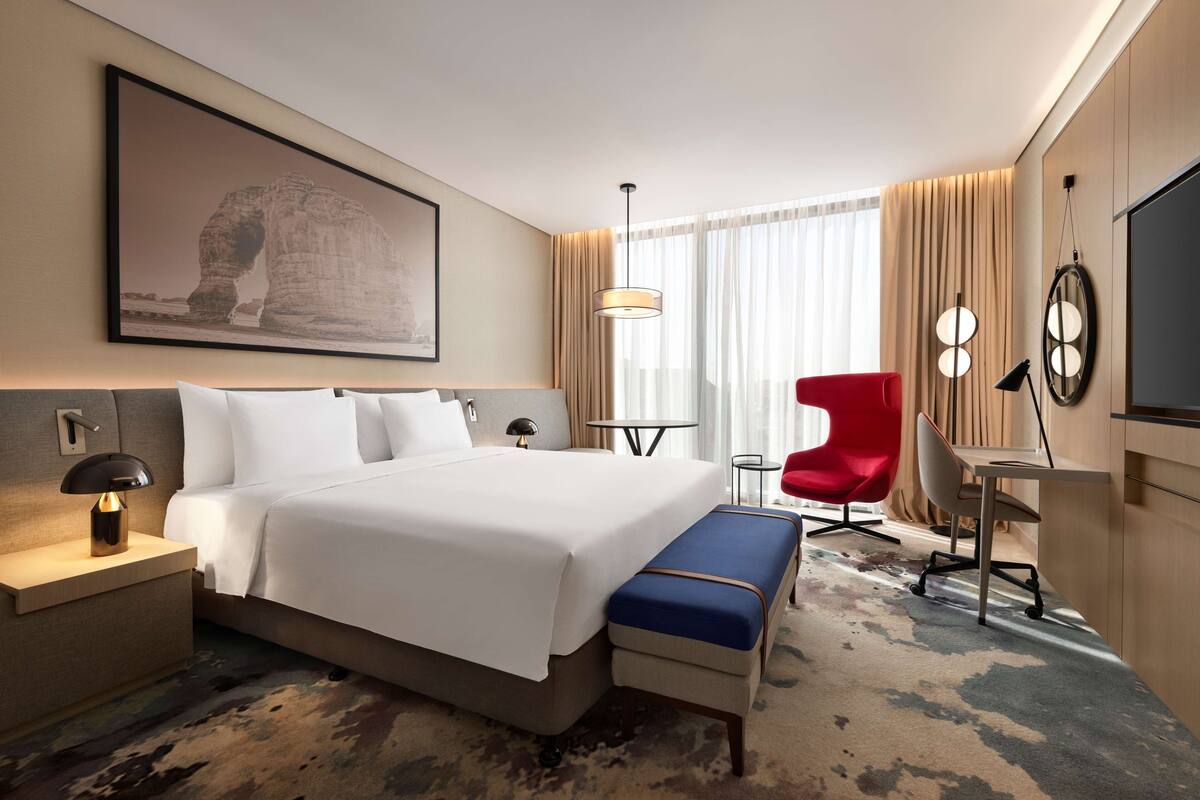 A hotel room with a large bed, a desk, a red chair, a small table, and a large window with curtains.