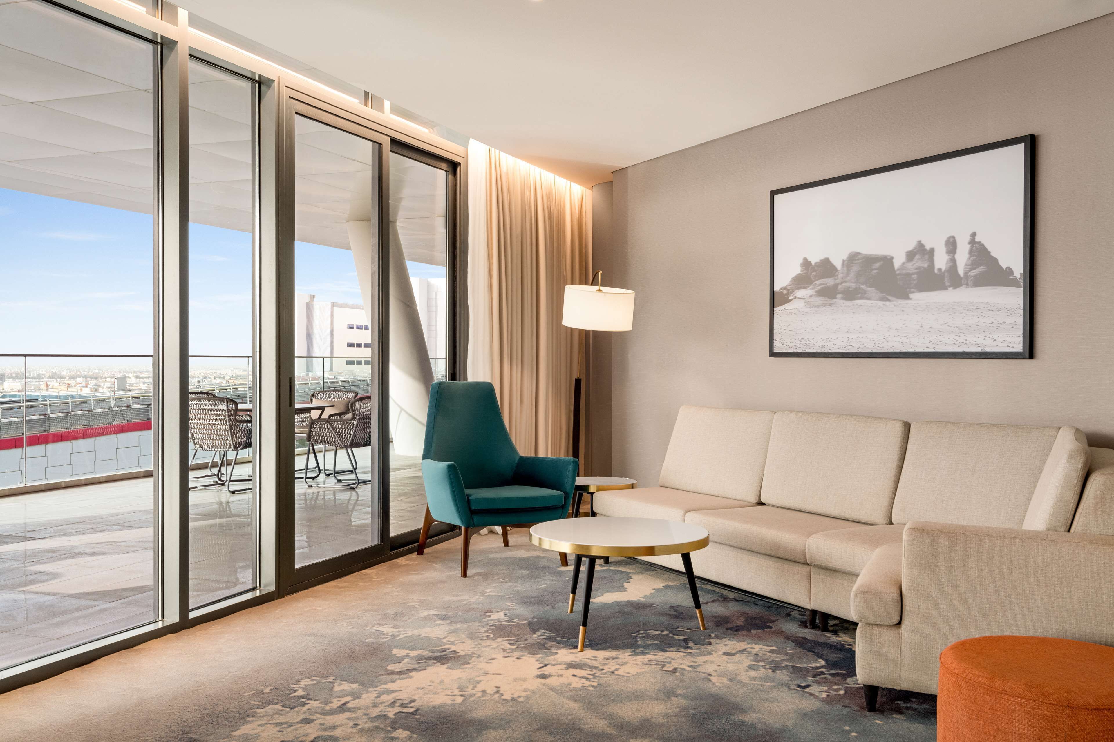 A modern hotel room with a large sliding glass door leading to a balcony, a beige sofa, a teal armchair, a small round table, and a floor lamp.