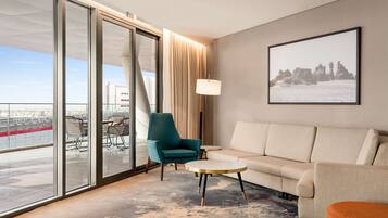 A modern hotel room with a large sliding glass door leading to a balcony, a beige sofa, a teal armchair, a small round table, and a floor lamp.