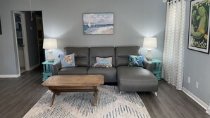 A living room with a gray sectional sofa, two side tables with lamps, a wooden coffee table, a patterned rug, a ceiling fan, a beach-themed painting, and a Florida-themed poster. - Family cottage by boat launch, minutes to Navarre or Pensacola Beach! (Gulf Breeze)