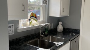 A kitchen with white cabinets, a black countertop, a stainless steel sink, and a window with patterned curtains. - Family cottage by boat launch, minutes to Navarre or Pensacola Beach! (Gulf Breeze)