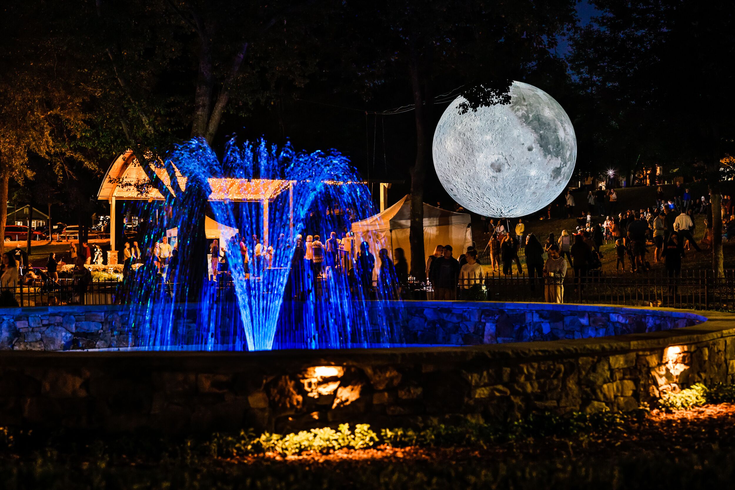 Escena nocturna con una fuente, una luna grande iluminada y una multitud de gente.