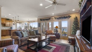 A spacious living room with a large sectional sofa, a coffee table, and a TV stand. There is a fireplace and a view of the outdoors through large windows.