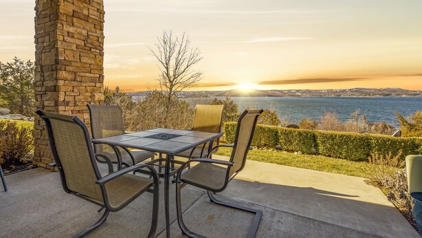 A patio with a stone pillar, a table, and chairs, overlooking a body of water and a distant cityscape during sunset.