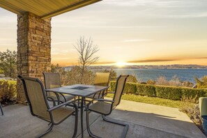 A patio with a stone pillar, a table, and chairs, overlooking a body of water and a distant cityscape during sunset.