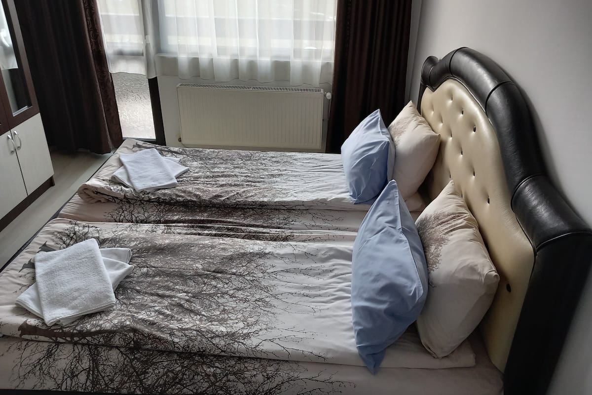 A hotel room with a bed, two pillows, a radiator, and a window with curtains.