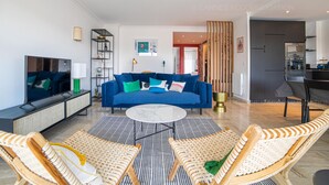 A modern living room with a blue sofa, wicker chairs, a TV, and a kitchen in the background.