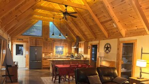 A spacious living area with a leather sofa, a dining table, and a kitchen with wooden cabinets and a large island. - Hill Country Ranch Retreat, Pool, Views, Sleeps 16+, Weddings & Family Groups (Florence)