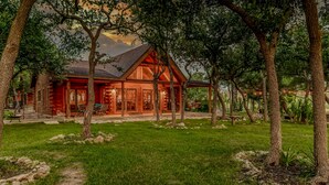 A log cabin with a porch, surrounded by trees and a grassy yard. - Hill Country Ranch Retreat, Pool, Views, Sleeps 16+, Weddings & Family Groups (Florence)