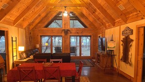 A spacious cabin with a high ceiling, a dining area with a red table and chairs, a living room with a sofa and chairs, and a large window offering a view of the outdoors. - Hill Country Ranch Retreat, Pool, Views, Sleeps 16+, Weddings & Family Groups (Florence)