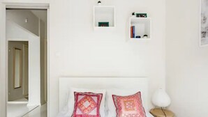 Un dormitori amb un llit blanc, dos coixins estampats, una petita taula de nit amb una làmpada i dues estanteries blanques amb llibres i objectes decoratius.