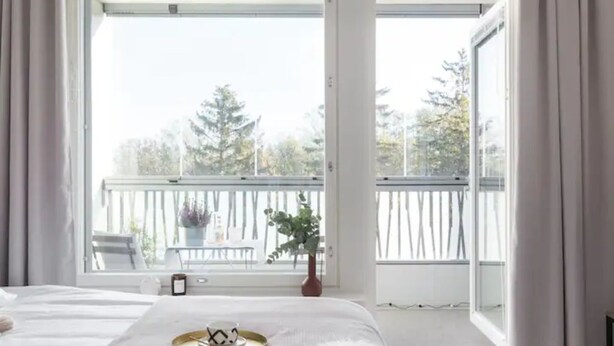 Ett sovrum med ett stort fönster, en säng med ett mjukt, vitt överkast och ett brickbord med en kopp och ett fat.
