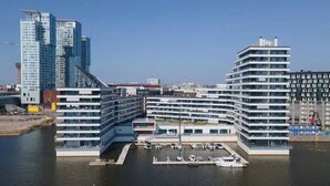 Un hotel frente al agua con varios pisos, un puerto deportivo con barcos y edificios modernos altos al fondo.