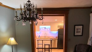 A living room with a sofa, armchair, and coffee table. There is a chandelier, a lamp, and a rug. A kitchen is visible through an open door. - Charming 3-bedroom house in lovely downtown Madison. (Madison)