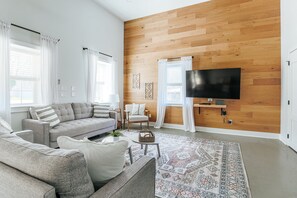A modern living room with a wooden wall, a grey sofa, a white armchair, a flat-screen TV, and a patterned rug.