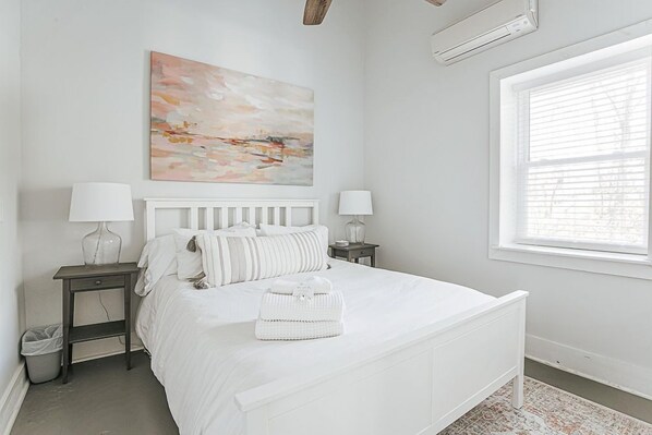 A white bedroom with a bed, two bedside tables, a nightstand, a window, and a painting on the wall.
