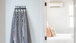 Ein Flur mit einem Handtuchhalter, der gestreifte Handtücher trägt, einem Bett mit weißer Matratze und Pfirsichkissen und einem Fenster mit Jalousien.