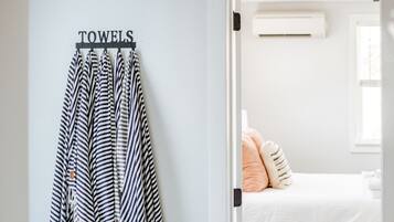 Un pasillo con un perchero que sostiene toallas a rayas, una cama con colchón blanco y almohadas color melocotón, y una ventana con persianas.