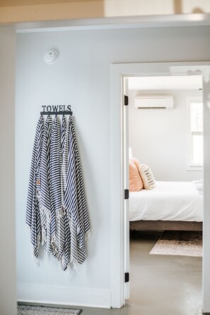 Un couloir avec un porte-serviettes où sont rangées des serviettes à rayures, un lit avec un matelas blanc et des oreillers couleur pêche, et une fenêtre avec des stores.