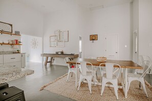 Une cuisine spacieuse avec des meubles blancs, une table à manger en bois et des chaises blanches. Un petit panier de basket est fixé au mur.