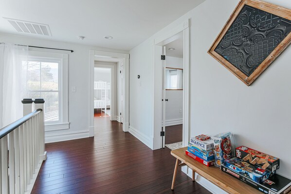 A hallway with wooden flooring, white walls, and a staircase with a white railing. There are boxes of board games on a table.