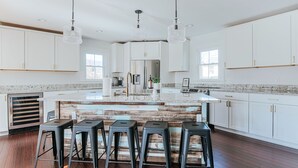 Cozinha moderna com armários brancos, uma ilha central com banquetas, eletrodomésticos de aço inoxidável e iluminação pendente.