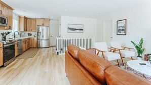 Et moderne køkken med træskabe, rustfrie hvidevarer og et spiseområde med en lædersofa og hvide stole.
