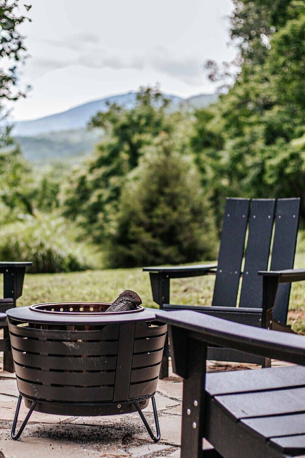 A black outdoor grill with a fire, surrounded by Adirondack chairs, set against a backdrop of greenery and mountains.