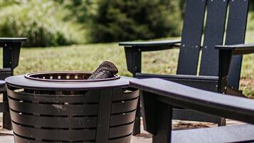 A black outdoor grill with a fire, surrounded by Adirondack chairs, set against a backdrop of greenery and mountains.