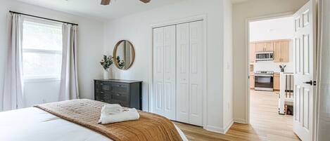 A bedroom with a bed, a ceiling fan, a dresser, a mirror, and a view into the kitchen.