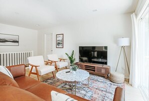 A modern living room with a brown leather sofa, white armchairs, a round coffee table, a large flat-screen TV, a floor lamp, and a patterned rug.