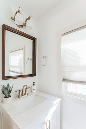 Une salle de bain avec un lavabo blanc, un miroir avec un cadre en bois, deux appliques murales et une fenêtre avec des stores.