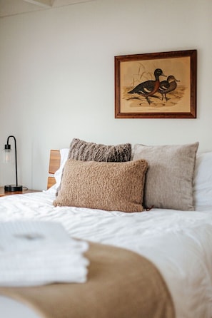A neatly made bed with two pillows and a blanket, a framed picture of birds on the wall, and a bedside table with a lamp.