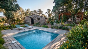 Une terrasse en bois avec une piscine, un bâtiment en pierre et une pergola dotée d’un espace salon.