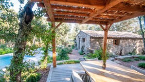 Une terrasse en bois avec une pergola, une maison en pierre et une piscine en arrière-plan.