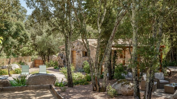 Un bâtiment rustique en pierre, entouré d’arbres, avec une terrasse en bois et un espace salon extérieur.