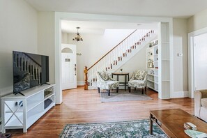 A living room with a wooden staircase, a television on a white stand, patterned chairs, and a bookshelf. - Oma's Garden Oasis: Spacious, Walk to Park (Staunton)